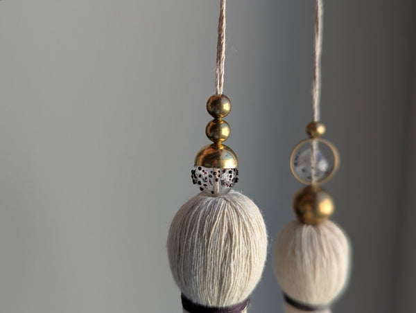 A close-up of a double tassel with deep purple colored thread, and glass and brass beads at the top against a neutral gray background
