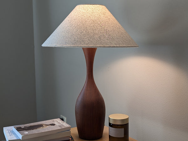 A freckled cotton lamp shade in an extreme tapered shape called a coolie shade on a vintage, teak wood lamp that sits on a nightstand with a candle, some books, and some jewelry