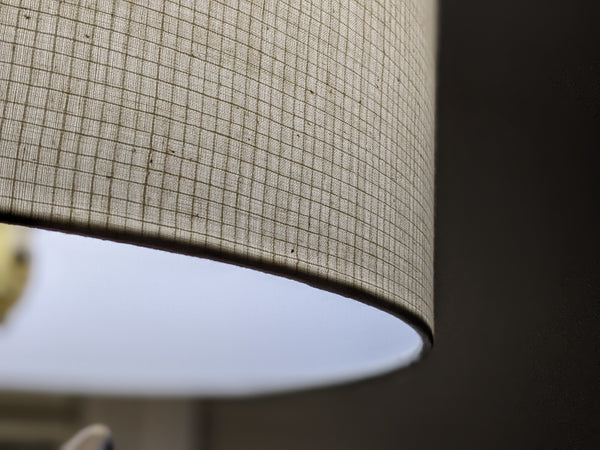 Close-up of a drum lampshade made of woven beige fabric with a subtle grid pattern, showing the underside rim and a soft white light glow inside.