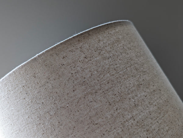 Speckled Oatmeal cotton fabric lampshade with brass hardware against a blurred background