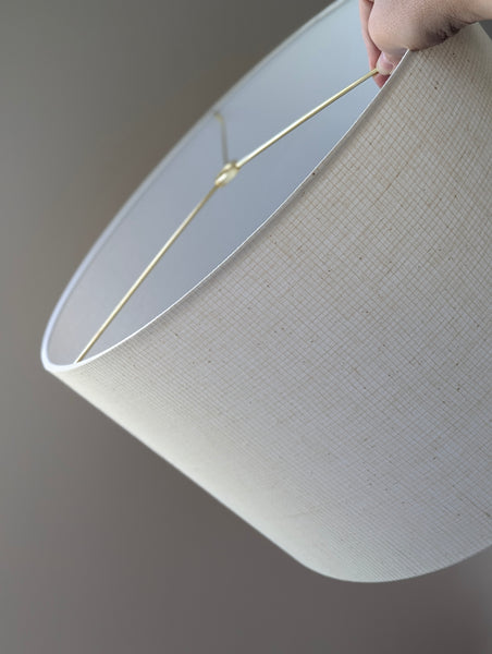 A close-up of a cream fabric lampshade being held by a hand, showing the interior brass spider washer and the textured fabric weave against a neutral gray background.