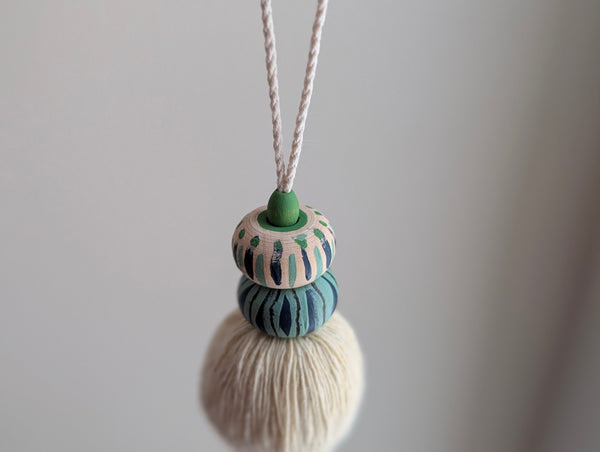 A close-up of a handmade wooden bead tassel, featuring a cream-colored cotton and blue and green accents against a soft beige background.