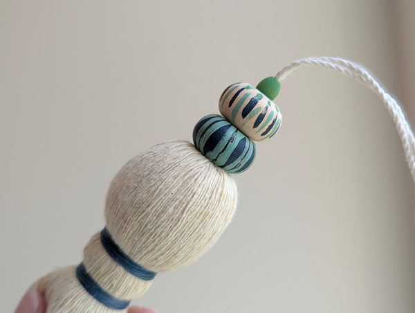 A close-up of a handmade wooden bead tassel, featuring a cream-colored cotton and blue and green accents against a soft beige background.