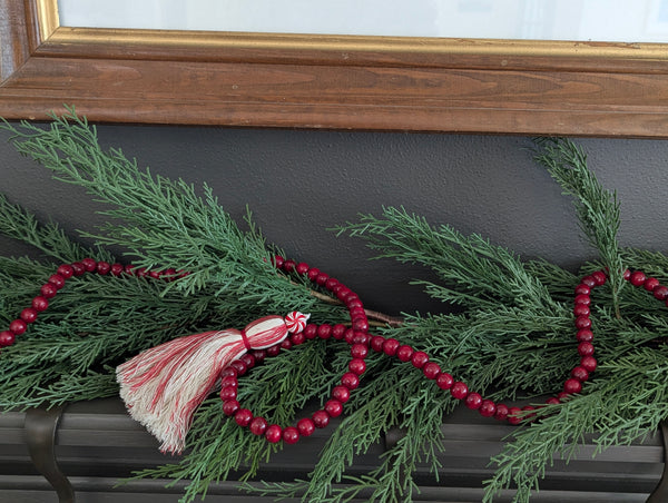 Red beaded garland with tassel on a green leafy branch against a dark wall.