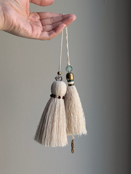 A hand holds two decorative cream-colored tassels with metallic and glass beads against a plain gray background.