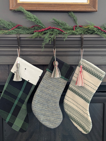 Three Christmas stockings hanging on a fireplace mantle decorated with greenery, garland, and holiday tassels.