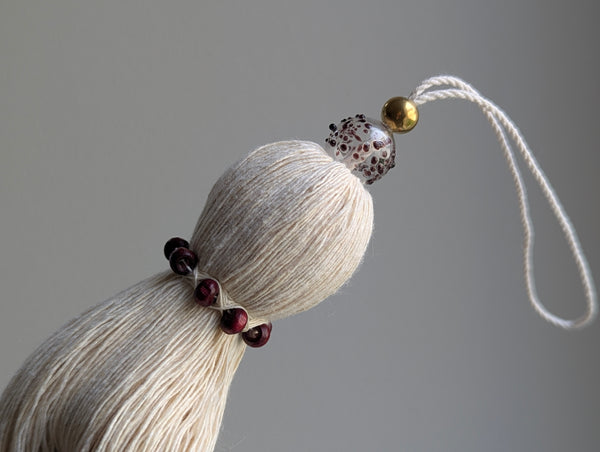 A close-up of a decorative tassel ornament made of cream-colored thread, accented with dark red beads, a clear glass bead with burgundy speckles, and a small gold bead, shown against a neutral gray background.