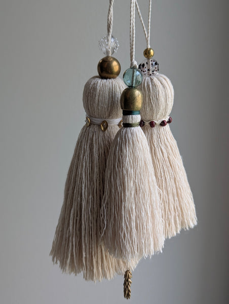 Three decorative hanging tassels made of cream-colored thread, embellished with brass-colored beads, a green glass bead, and small dark red beads, suspended against a neutral gray background.