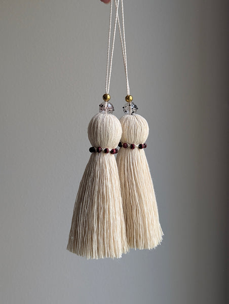 Two cream-colored decorative tassels hanging from white cords with small gold and burgundy glass bead accents, set against a plain gray background.