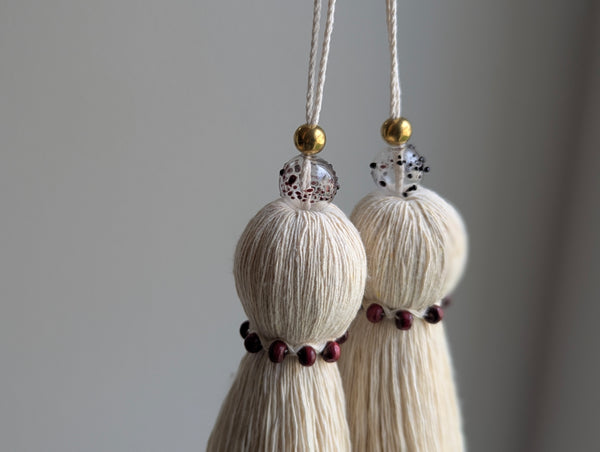 Two cream-colored cotton tassels with gold and red speckled beads hanging against a neutral gray background.