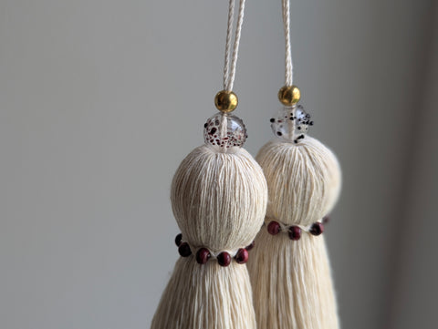 Two cream-colored cotton tassels with gold and red speckled beads hanging against a neutral gray background.