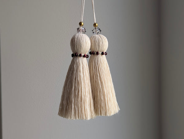 Two cream-colored cotton tassels hanging from white cords with small red beads and clear crystal and gold accents, set against a plain gray wall.