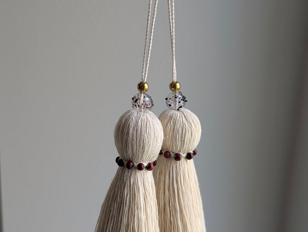 Two cream-colored cotton tassels hanging from white cords with gold beads and small dark red accents, set against a soft gray background.