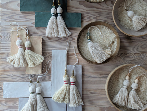 Twelve decorative tassels made of off-white cotton thread, each featuring a loop for hanging. The tassels are adorned with various accents including gold-toned metal beads, clear glass beads, and colored thread wraps in pink, green, and red.