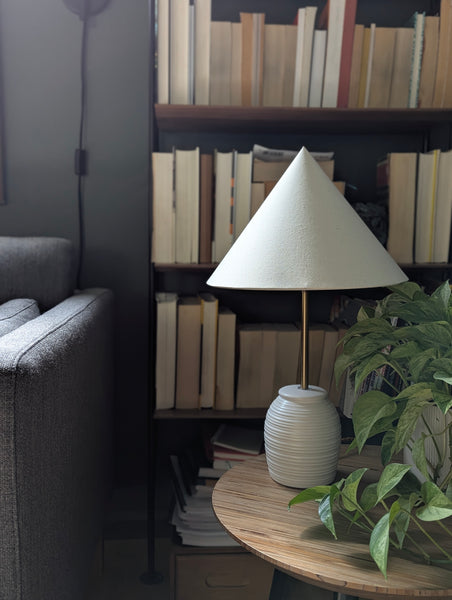 cone shaped lamp shade on a white ceramic table lamp in a living room with a bookcase in the background and a plant on the side table