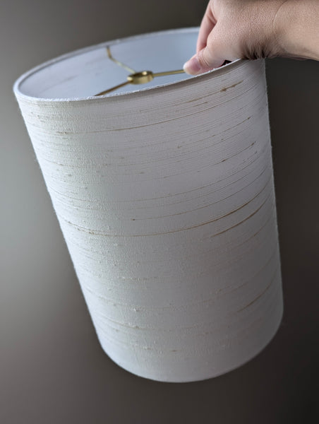 A hand holding a white drum lampshade with brass spider washer, shown against a dark gray background.