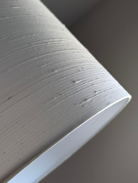 Close-up of a drum lampshade with a textured off-white surface and a smooth curved edge, photographed against a soft gray background.
