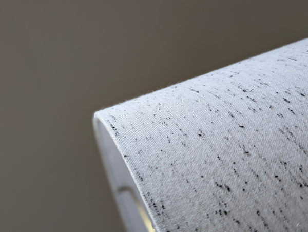 close up of a hand holding a white lamp shade with small black flecks throughout the fabric resembling a cookies and cream milkshake