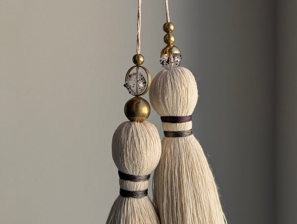 A close-up of a double tassel with deep purple colored thread, and glass and brass beads at the top against a neutral gray background