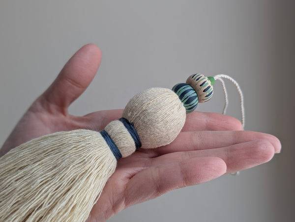 A close-up of a handmade wooden bead tassel, featuring a cream-colored cotton and blue and green accents against a soft beige background.