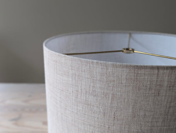 Close-up of a tan and gray linen lampshade with brass hardware against a blurred background