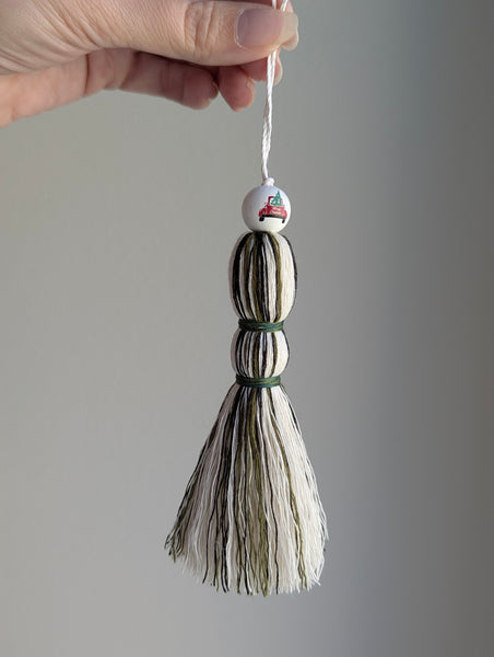 Holiday tassel ornament in green, white, and black with a top bead featuring a red truck carrying a Christmas tree.