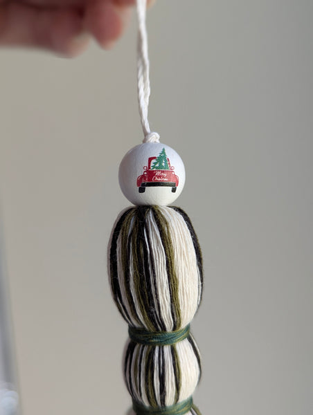 Holiday tassel ornament in green, white, and black with a top bead featuring a red truck carrying a Christmas tree.