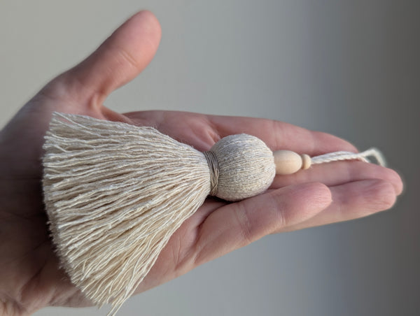 A hand holding a cream-colored tassel ornament with a textured spherical bead and small wooden beads on a cord, set against a soft gray background.