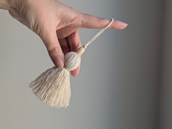 A hand holding a small decorative tassel with a round wooden bead and a light beige cord against a soft gray background.