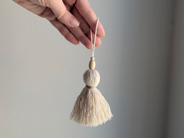 A hand holding a small decorative tassel ornament with a light wood bead and a cream-colored thread tassel against a plain gray background.