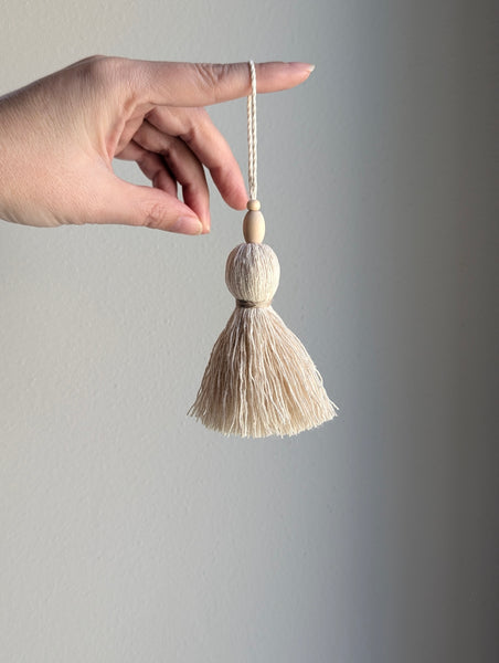 A hand holding a small decorative tassel ornament against a plain gray background. The ornament has a cream-colored cotton tassel, two round wooden beads, and a matching cream cord.
