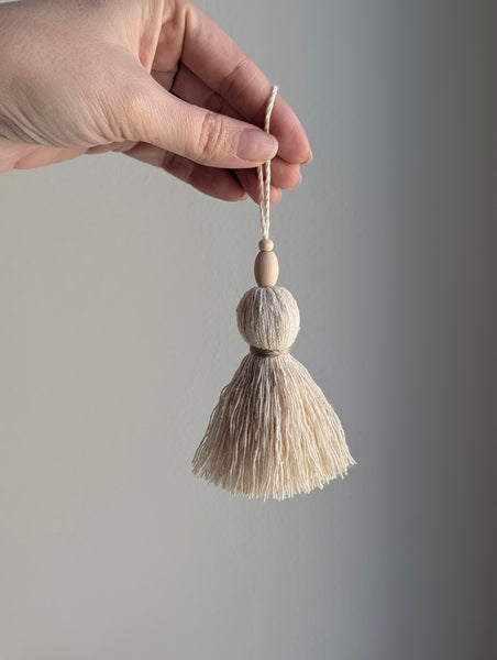 A hand holding a small decorative tassel ornament with two wooden beads and a cream-colored cord, set against a plain gray background.