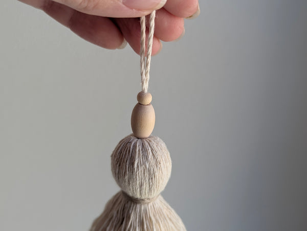 A close-up of a hand holding a light beige tassel ornament with two wooden beads and a white hanging string against a soft gray background.