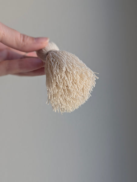 A hand holding a small, soft-textured cream-colored brush against a plain gray background.