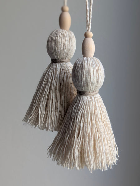 Two hanging tassel ornaments with natural wood beads and cream-colored fringed tassels, shown against a soft gray background.