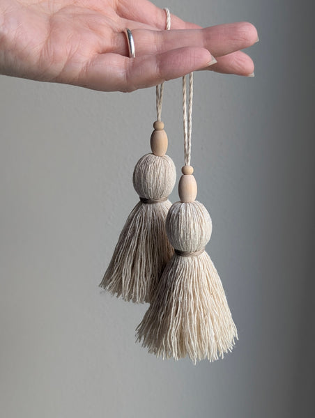 A hand holding two decorative tassel ornaments made of light beige cord and small wooden beads, shown against a plain gray background.