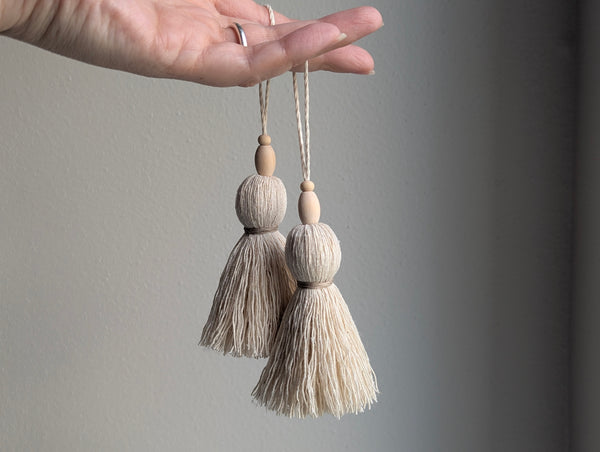 A hand holding two decorative tassel ornaments with light wood beads and cream-colored threads against a plain gray background.