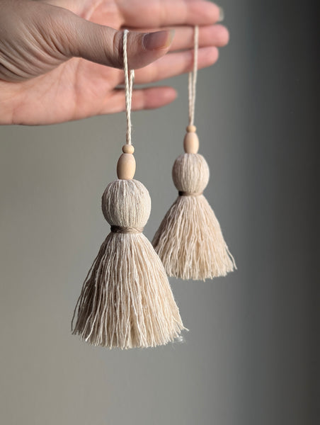 A close-up of a hand holding two cream-colored cotton tassel ornaments with wooden beads and hanging loops against a soft gray background.