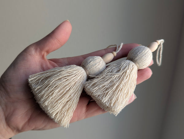 A hand holding two cream-colored tassel car keychains with wooden beads against a neutral gray background.