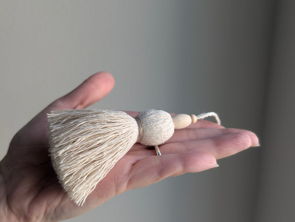 A hand holding a small decorative tassel ornament made of cream-colored thread, a round bead, and a light wood bead, against a soft gray background.