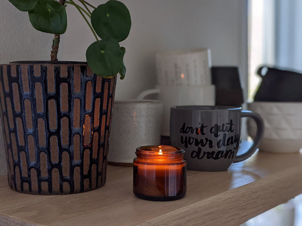 an amber glass candle with the label removed on a white oak self with coffee mugs and a plant