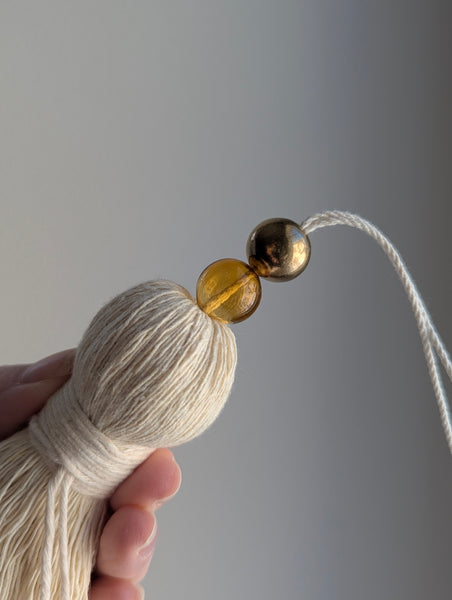 Tassel with amber glass bead and brass bead held by a hand against a gray background
