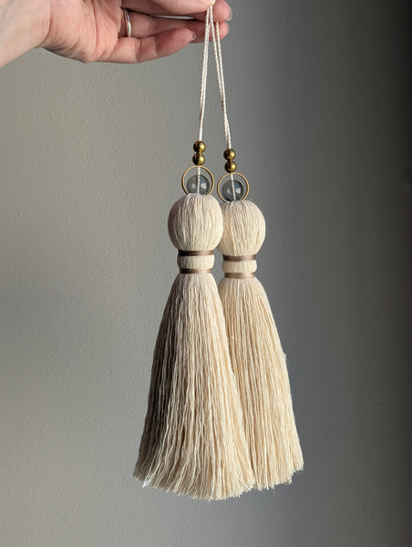 A close-up of two modern tassels with taupe colored thread, a gray glass bead and brass beads shown against a soft gray background.