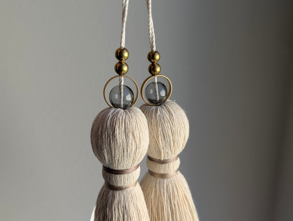 A close-up of two modern tassels with taupe colored thread, a gray glass bead and brass beads shown against a soft gray background.