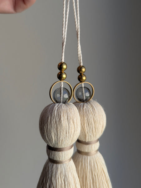 A close-up of two modern tassels with taupe colored thread, a gray glass bead and brass beads shown against a soft gray background.