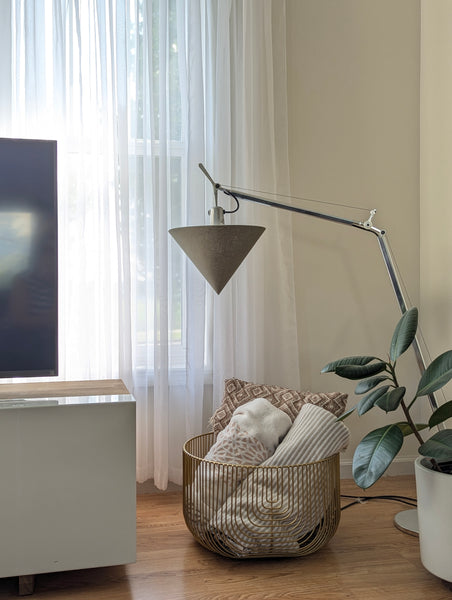 Modern living room with light wood floors, warm white walls, a basket of blankets and pillows, a plant, and a tv stand, and in the center, a small arching floor lamp with a green linen cone lamp shade