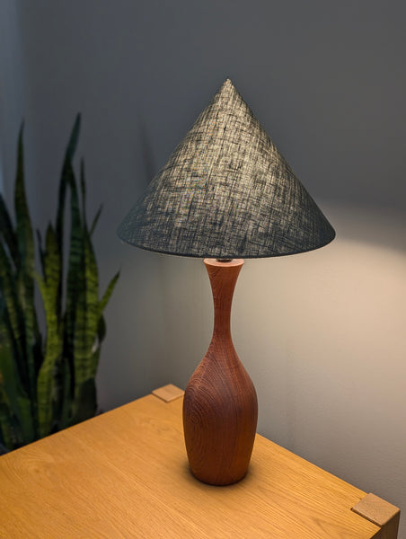 vintage walnut table lamp with a lit, cone lamp shade in green on a wooden nightstand next to a plant