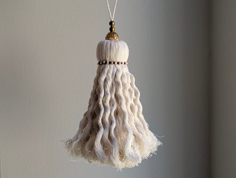 A close-up of a door knob tassel with deep purple colored thread, and wooden and brass beads at the top against a neutral gray background