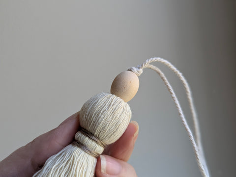 A close-up of a decorative tassel with cream-colored thread, a tan wrapped band, and a wooden beads shown against a neutral gray background