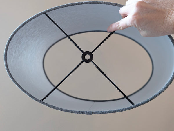 looking straight through the center of an oval shaped lamp shade made of grey fabric with black spider ring hardware